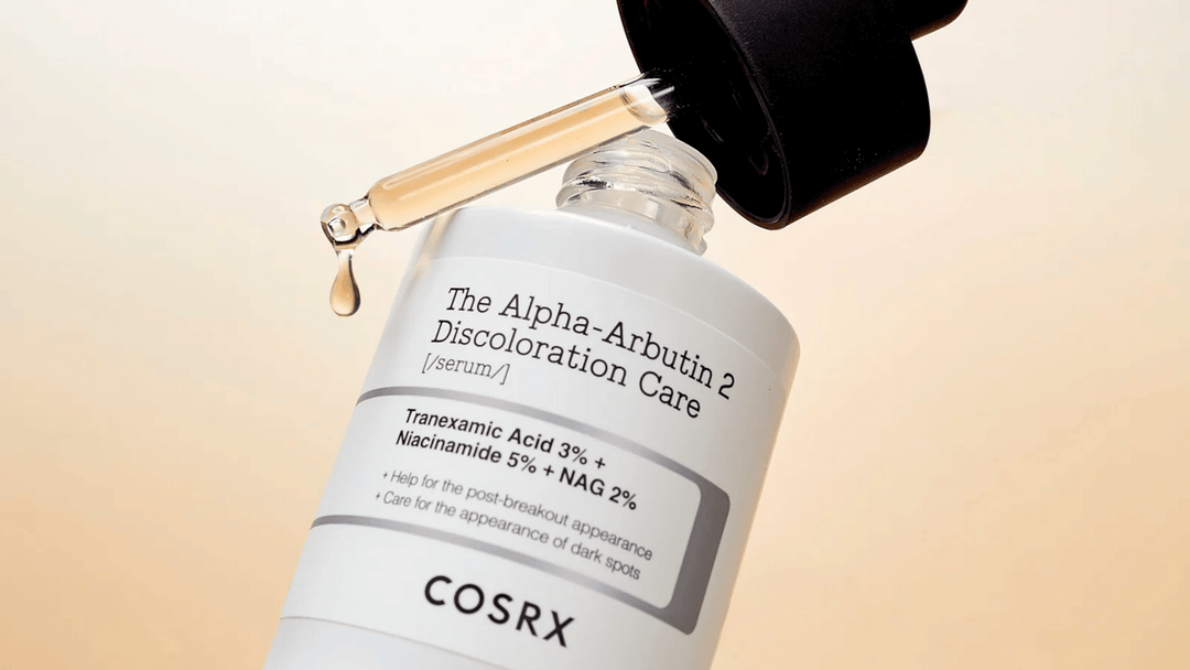 Pipetten droppar serum från en COSRX-flaska med texten "The Alpha-Arbutin 2 Discoloration Care". Produkten innehåller tranexamsyra och niacinamid för att behandla hyperpigmentering och mörka fläckar.