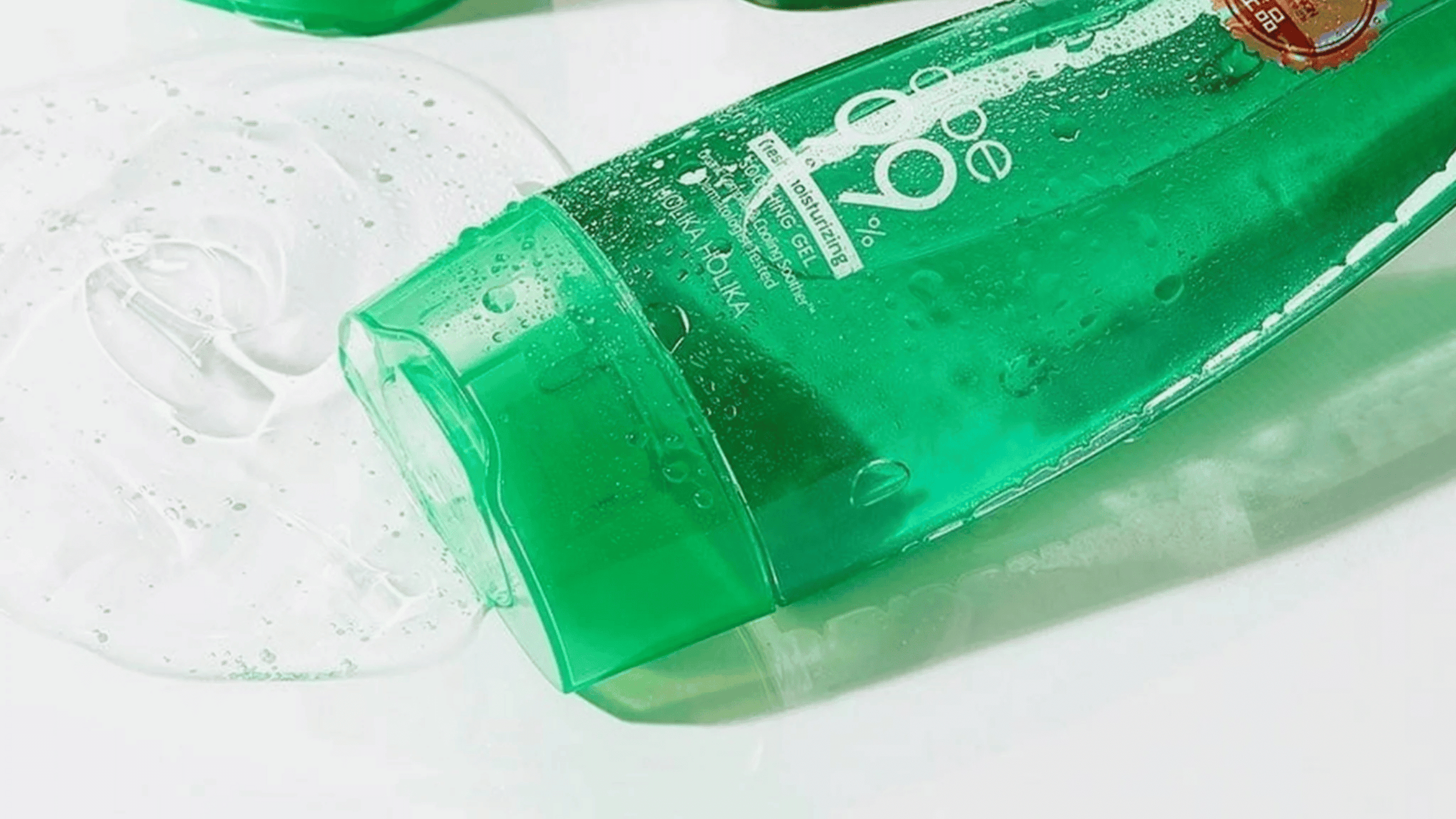 En grön tub med Holika Holika Aloe 99% Soothing Gel ligger på sidan med gelén utspridd bredvid. Tuben är delvis täckt av vatten och visar text som beskriver produktens återfuktande och lugnande egenskaper.