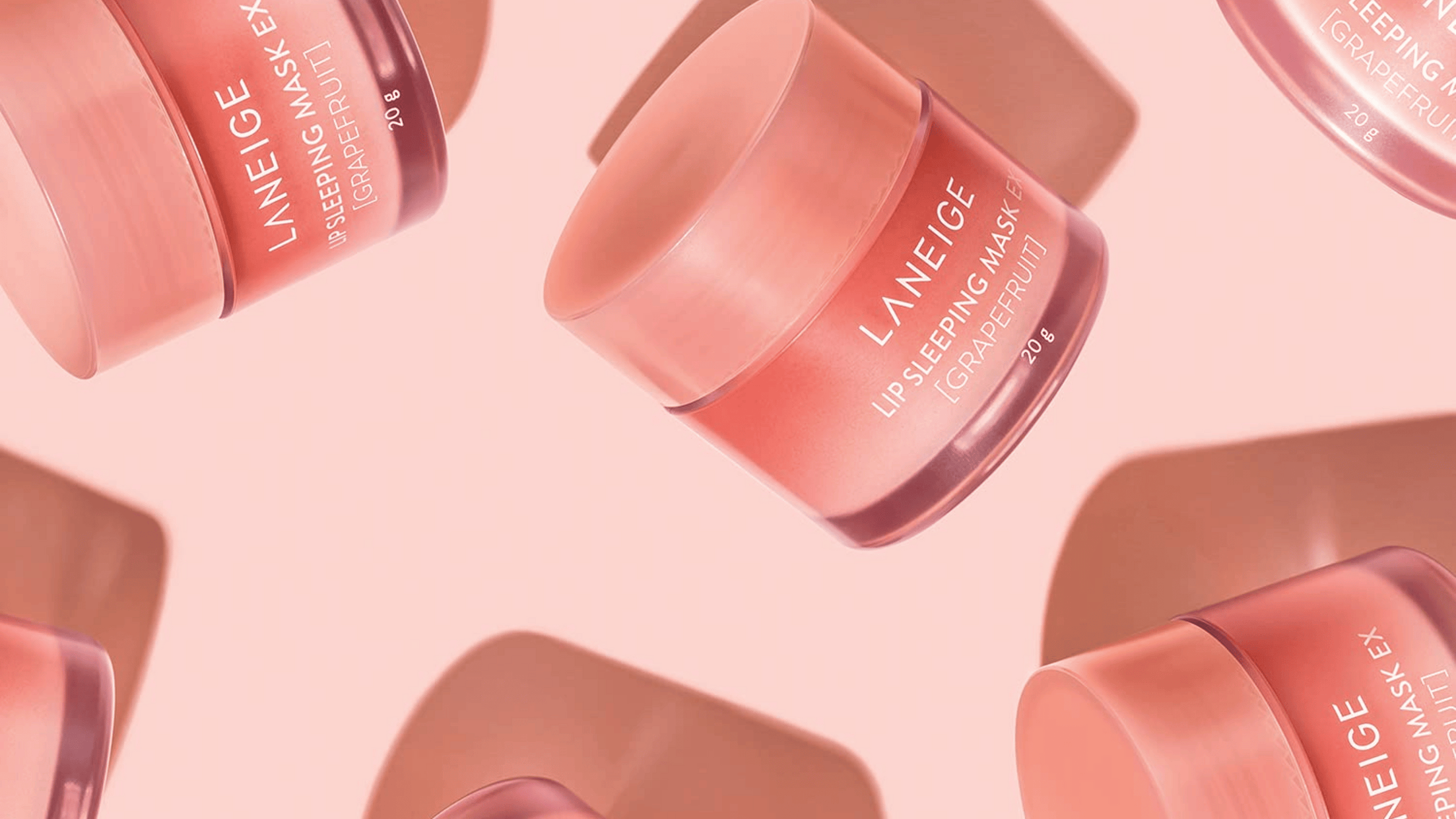 Flera genomskinliga rosa burkar av LANEIGE Lip Sleeping Mask i grapefrukt, spridda på en rosa bakgrund.