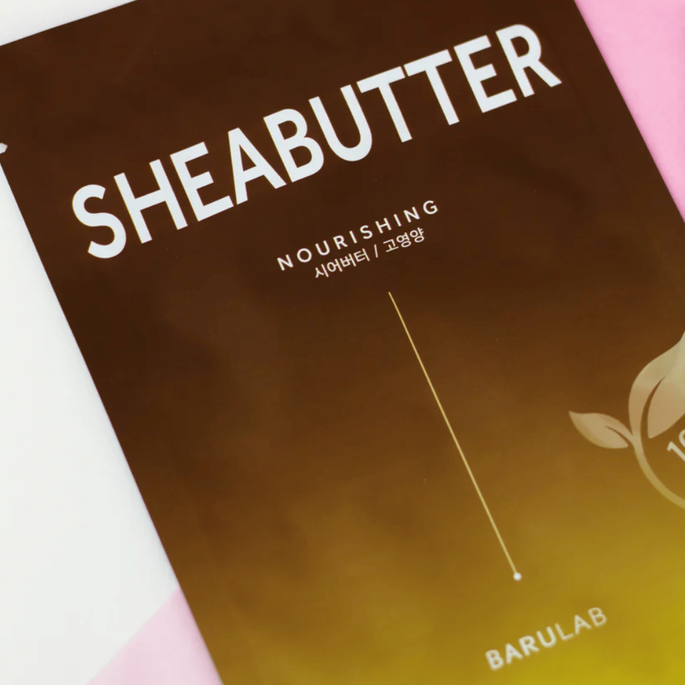 En närbild av en förpackning av BARULAB "SHEABUTTER Nourishing" mask. Produkten är märkt som "100% Vegan Product" och har en färggradient från mörkbrun till ljusbrun.