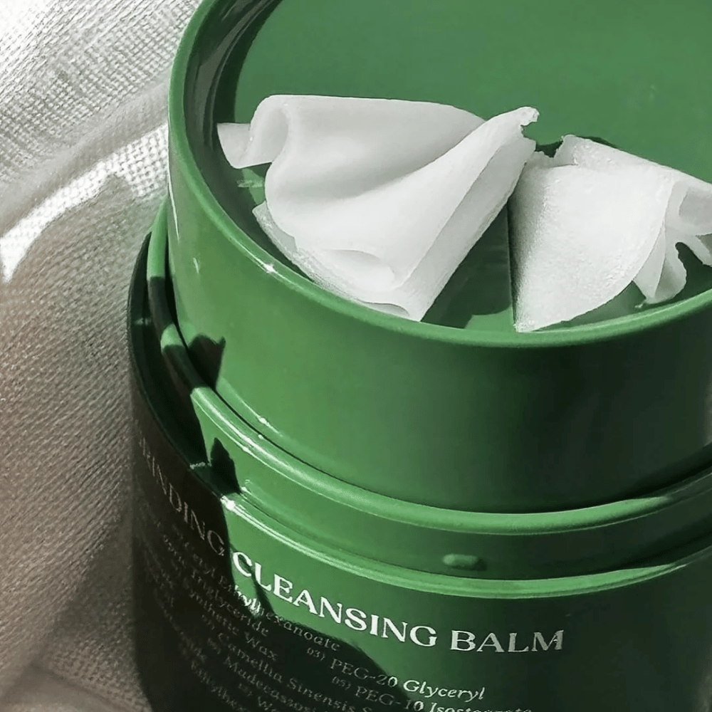 Bilden visar en grön burk med "Cleansing Balm", som är öppnad för att avslöja den medföljande spateln och det mjuka innehållet. Den gräddiga konsistensen reflekterar ljuset, vilket ger en känsla av lyx och effektivitet i rengöringen.