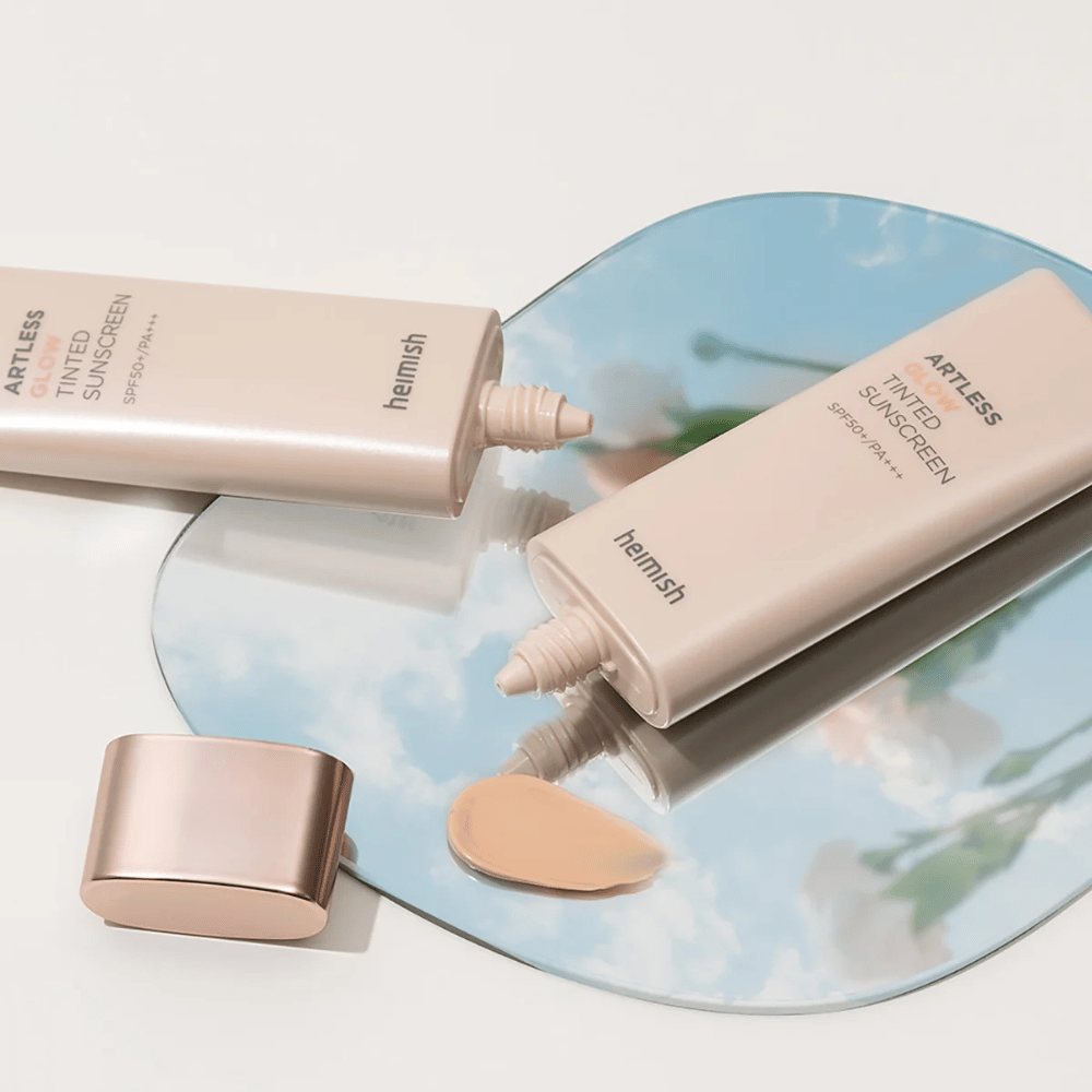 En flaska heimish Artless Glow Tinted Sunscreen SPF50+ PA+++ 40ml och en flaska concealer på en spegel, som visar olika hudtoner.